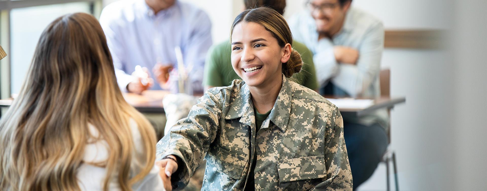 Photo of a young woman in a camo uniform smiling and shaking another young woman's hand