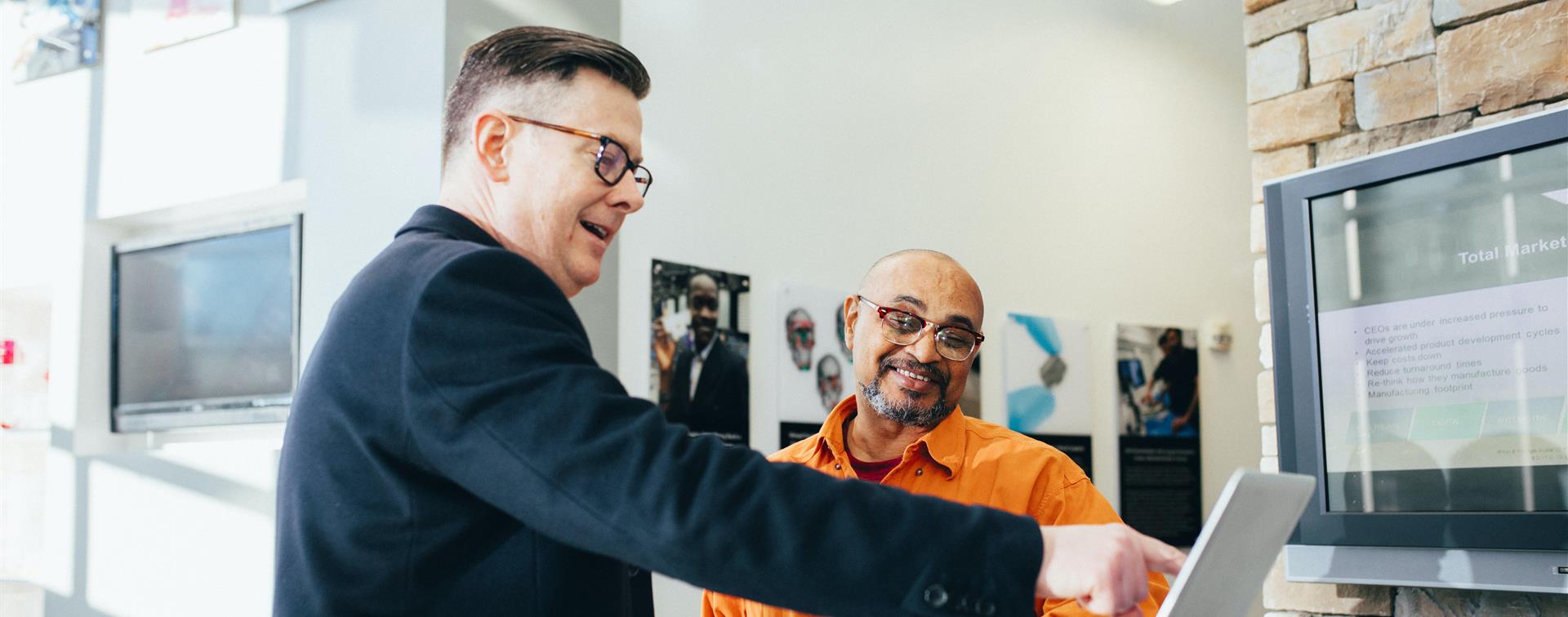 Two men stand infront of a computer wearing buisness attire. One also wears a bright orange shirt.