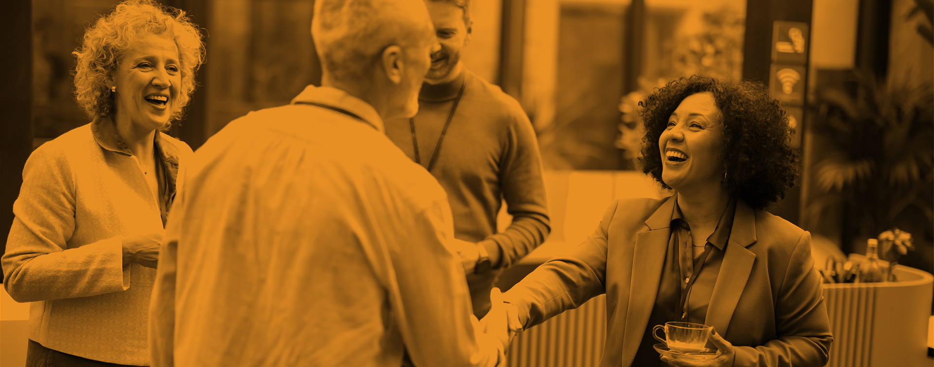 A woman in the center of the image shakes hand with a man in a conference like setting with others. There is an orange overlay.