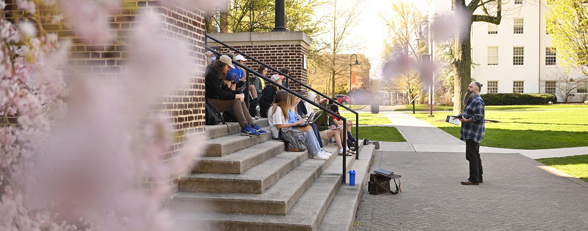 Photo of a professor addressing a group of college students meeting outside on the steps of an academic building on a sunny day
