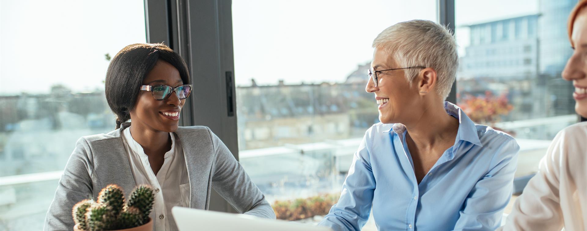 Photo of two professional women talking at a table with an open laptop