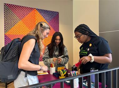 Three students stand around a portable cart with button-making materials on it and make voting-themed buttons.