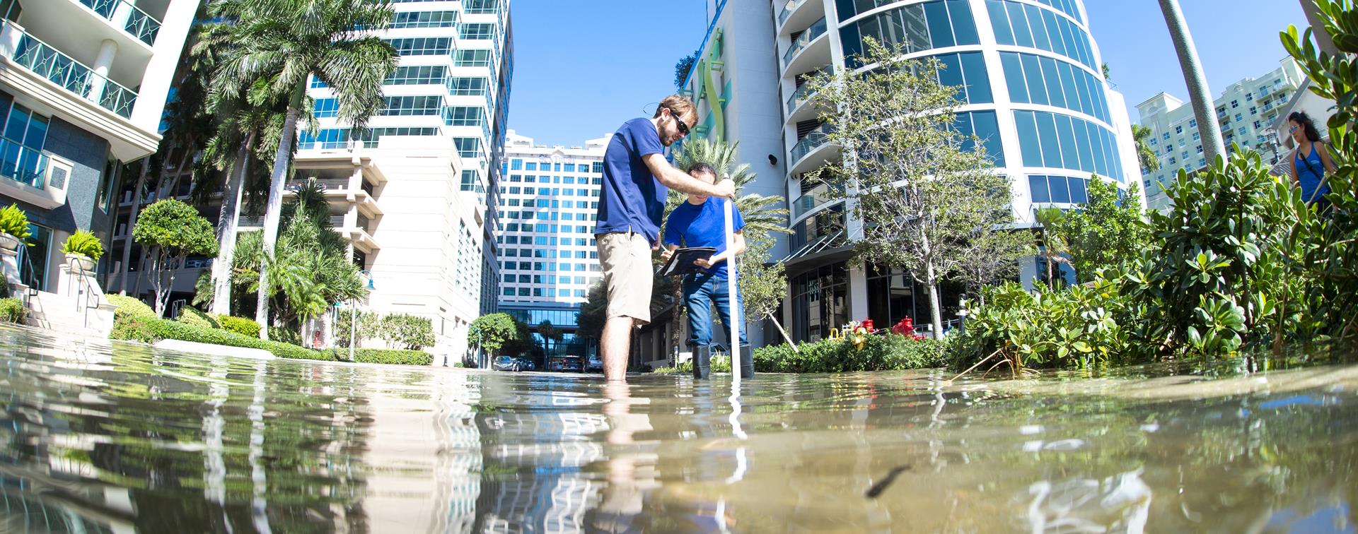 Photo of two students in ankle-deep water in a built-up area. They're holding a tube and studying the water.