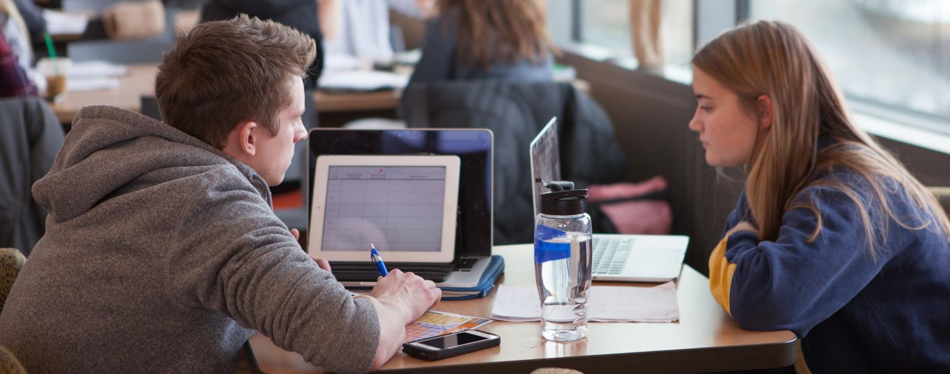 Photo of two college students sitting at a wooden table and studying on their laptops