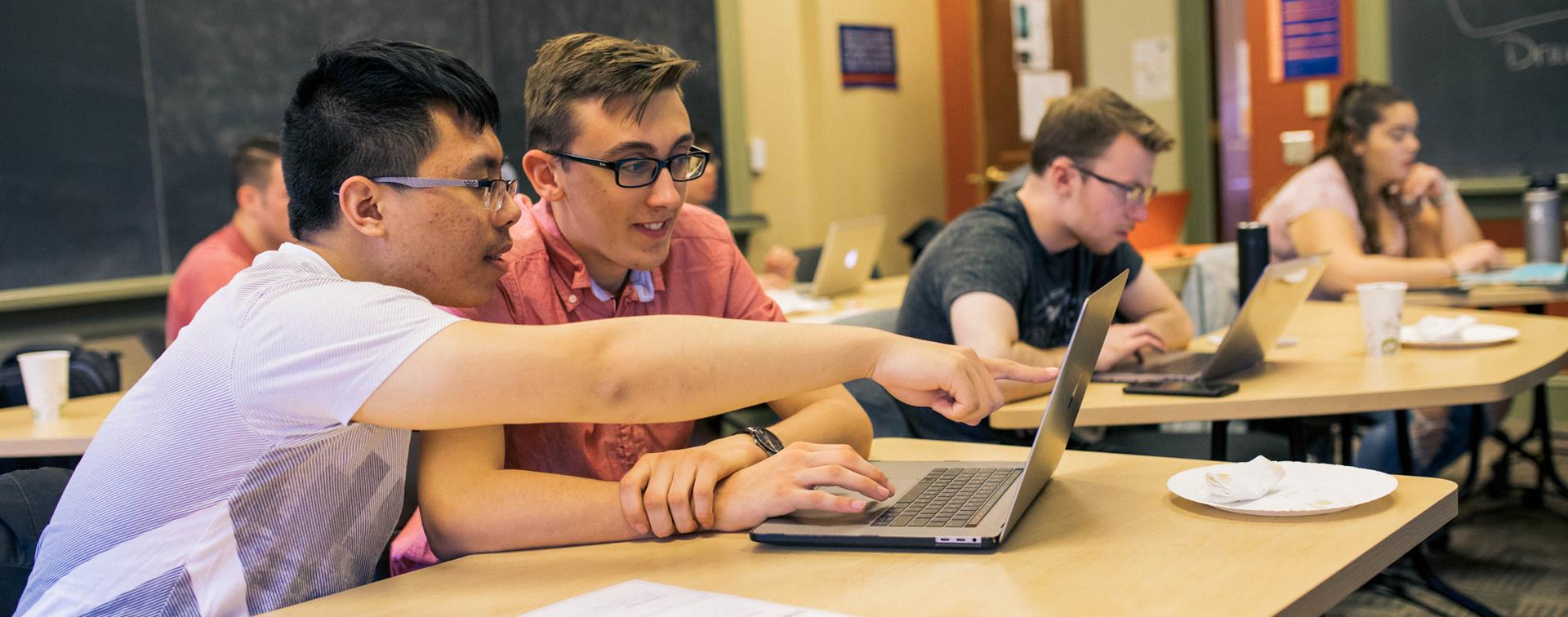 Photo of two college students sitting at a desk in a classroom. One is pointing at something on the other's laptop screen.