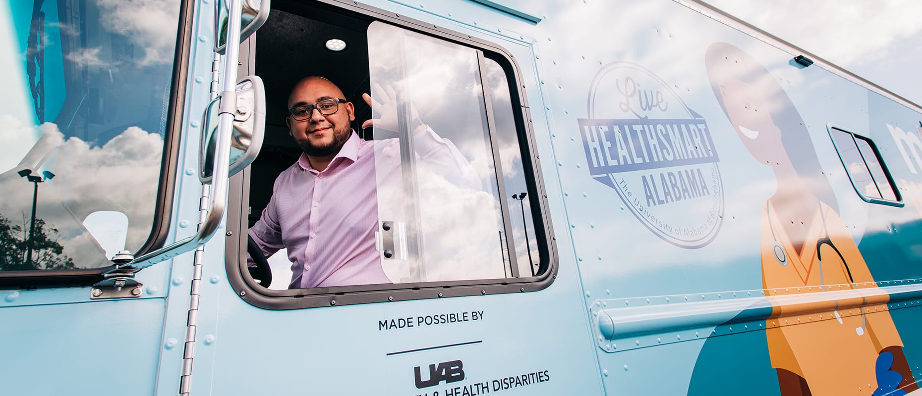 Photo of a man waving from the driver's seat of a van. The van is light blue and has a picture of a woman wearing medical gear.